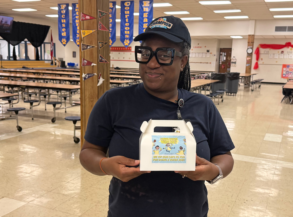 A cafeteria worker wearing glasses and a “Champion” cap holds a small box labeled “School Nutrition Hero Day” in a school cafeteria.