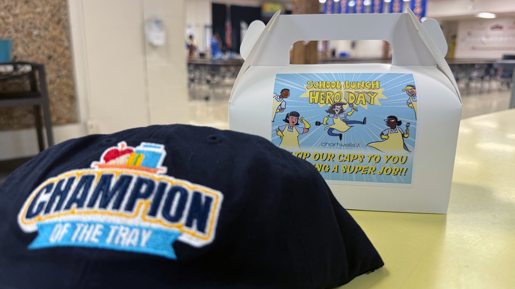 A navy cap embroidered with “Champion of the Tray” sits in front of a box displaying a colorful School Lunch Hero Day sign.