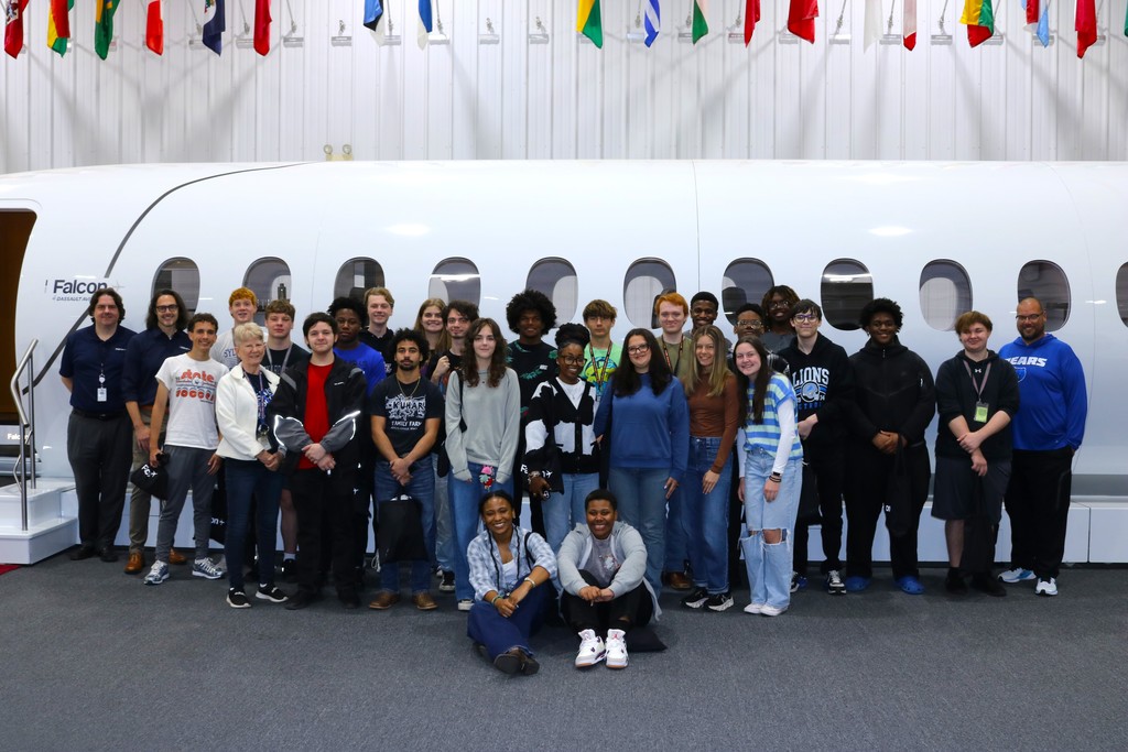 A large group of students and adults pose in front of a white aircraft fuselage labeled “Falcon” inside an aviation facility with international flags overhead.