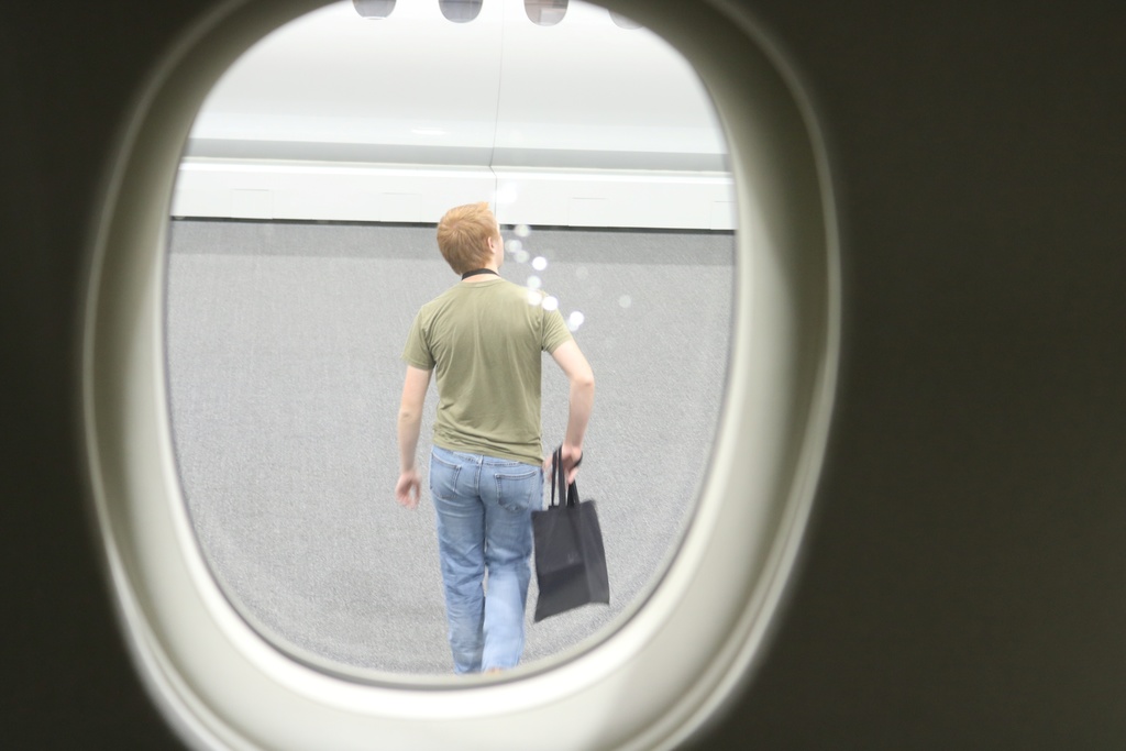 Seen through an airplane window, a person in a green shirt walks across the showroom floor carrying a black tote bag.
