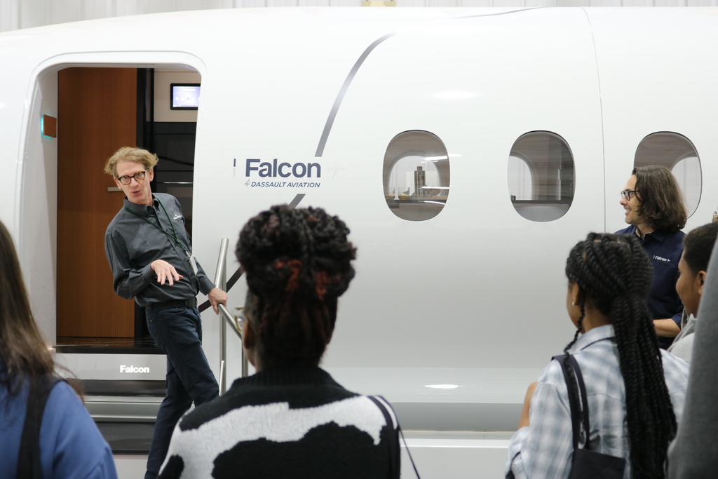 A person stands at the entrance of a Falcon aircraft explaining features to a group gathered in front of the fuselage.