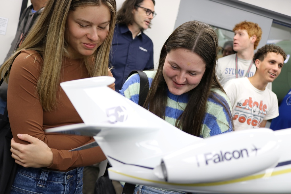A group gathers around a detailed model jet labeled “Falcon,” with two students closely observing the aircraft.