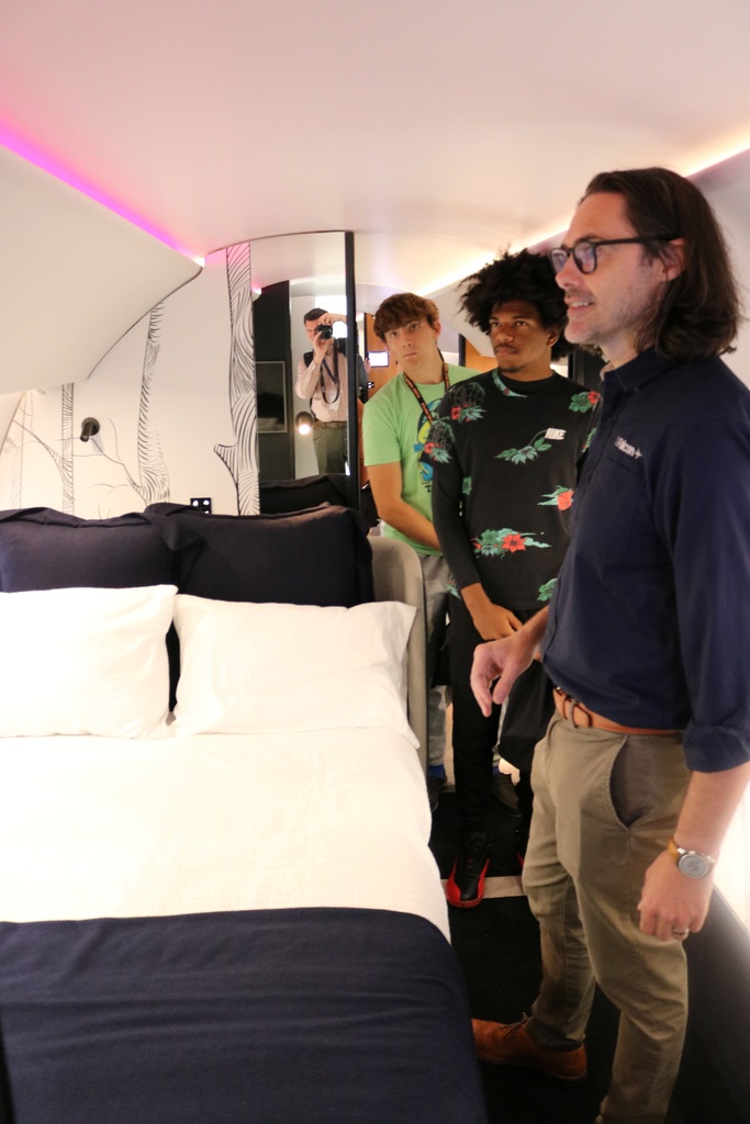 Four people stand inside a small, modern room with a neatly made bed, curved ceiling lighting, and wall art; one person photographs the space using a camera.