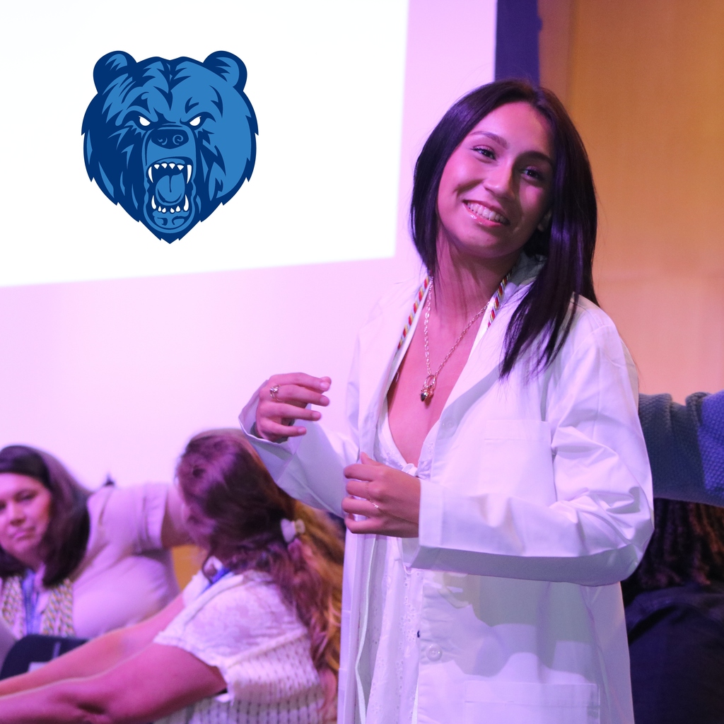 A student puts on a lab coat during a cording ceremony. Sylvan Hills High School logo is present.