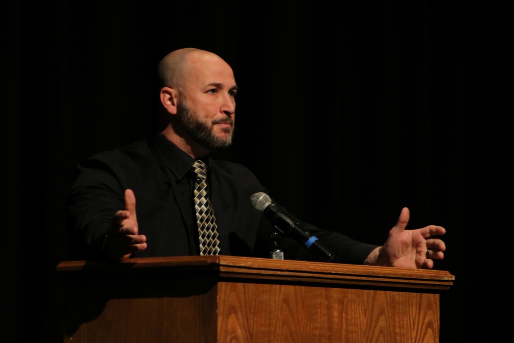 A presenter in a black shirt and patterned tie speaks from a wooden podium, gesturing with both hands on a darkened stage.