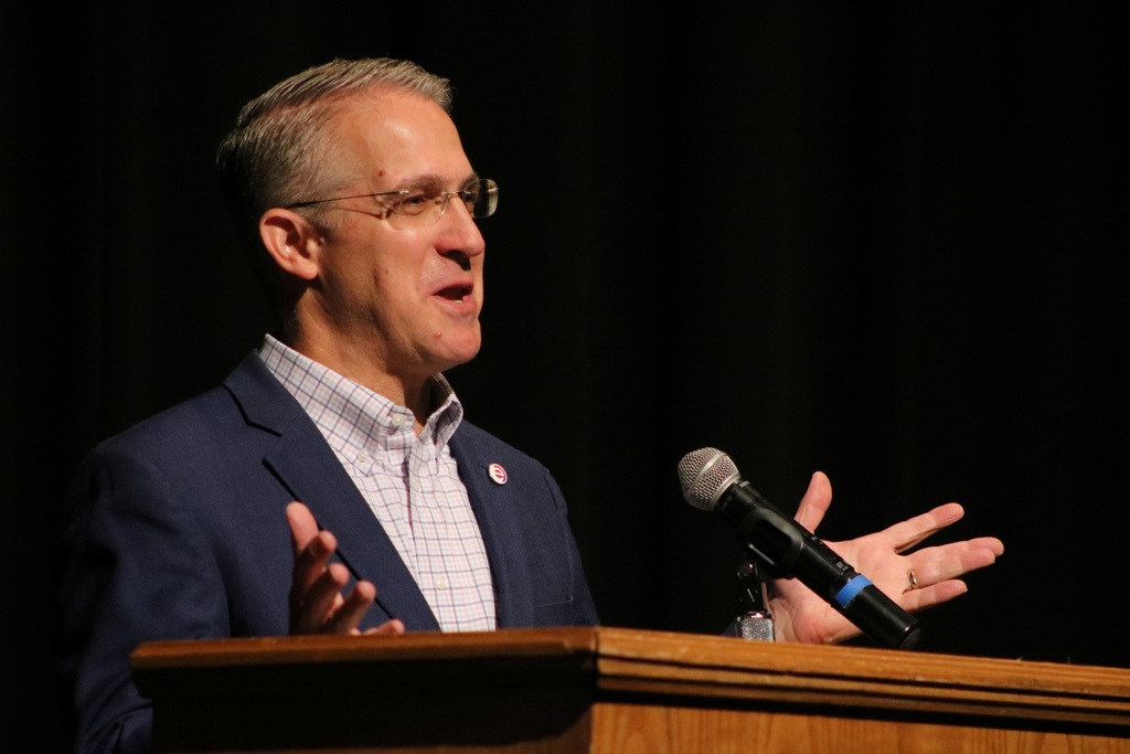 A presenter in a blue suit jacket and glasses speaks at a wooden podium, raising both hands while addressing the audience on a dark stage.
