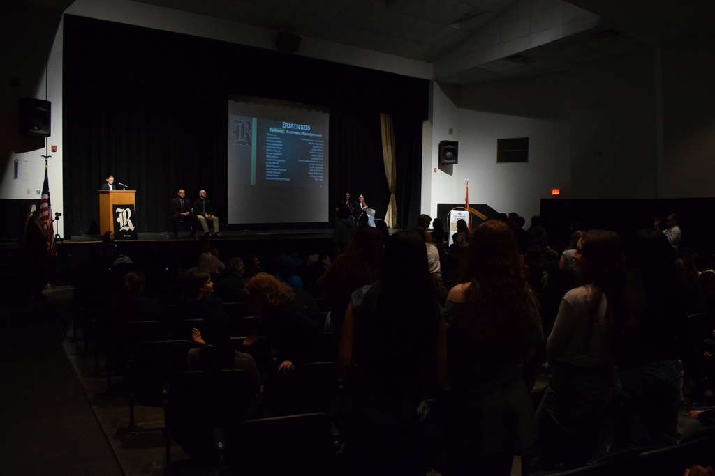 An auditorium audience faces a stage where a speaker stands at a podium. A large screen displays a slide titled “BUSINESS” with a list of names beneath it.