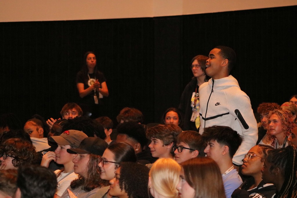 A packed auditorium of students sits facing forward. One student in a white Nike jacket stands among the crowd, and two adults stand near the back wall.
