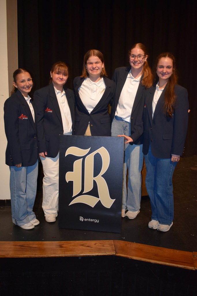 Five students stand on a stage in navy blazers and jeans behind a large Entergy‑branded sign with a stylized “R.” They face the audience in front of a black curtain during the Pledge to Graduate ceremony.