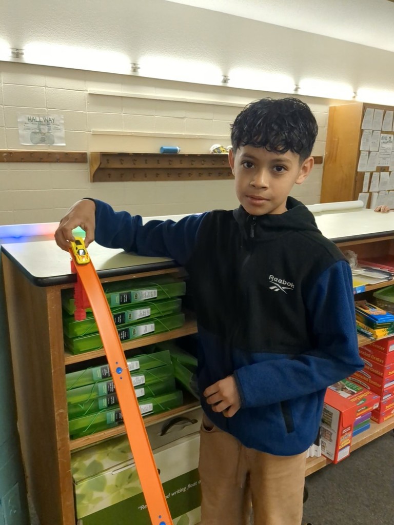 A student stands beside a classroom shelf, holding a yellow toy car at the top of an orange track angled downward. Classroom supplies and bulletin board papers are visible behind them.