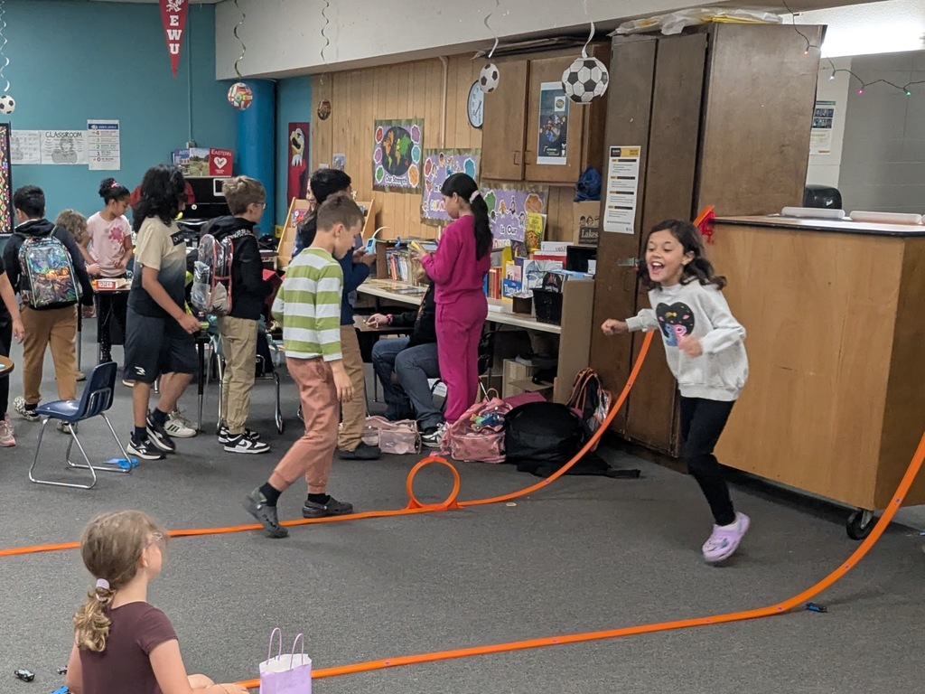 Children move around a classroom during a hands‑on activity. One child runs near an orange looping toy racetrack set up on the floor while others gather backpacks or play at stations.