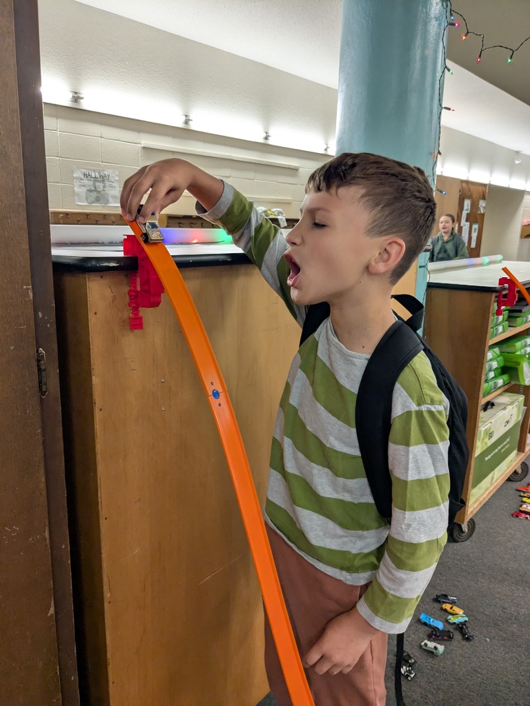 A child holds a toy car at the top of an orange ramp attached to a tall wooden surface, preparing to release it. Several toy cars are scattered on the floor below.