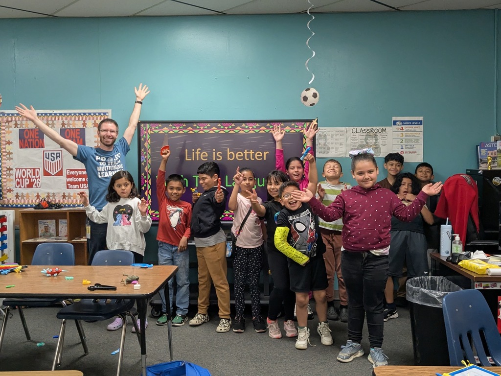 A group of children and an adult stand together in a classroom, smiling with their arms raised. The adult’s shirt reads “Proud to Teach Multilingual Learners,” and posters decorate the teal walls.