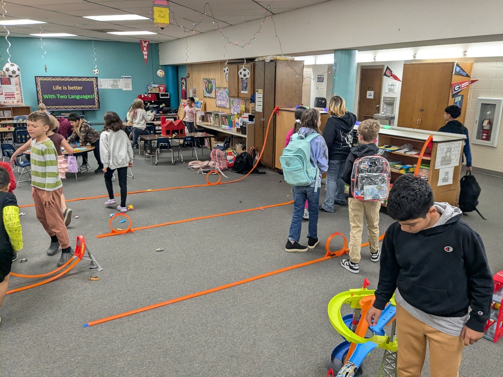 Students in a classroom experiment with several orange toy car tracks arranged on the carpet. Cars race through loops and ramps while children observe and test different setups.