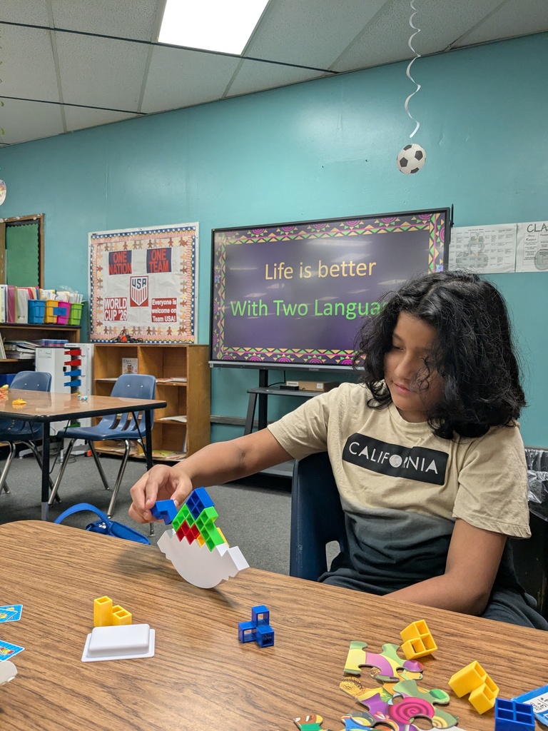 A student sits at a table assembling colorful interlocking blocks and puzzle pieces. Behind them, a classroom display reads “Life is better with two languages.”