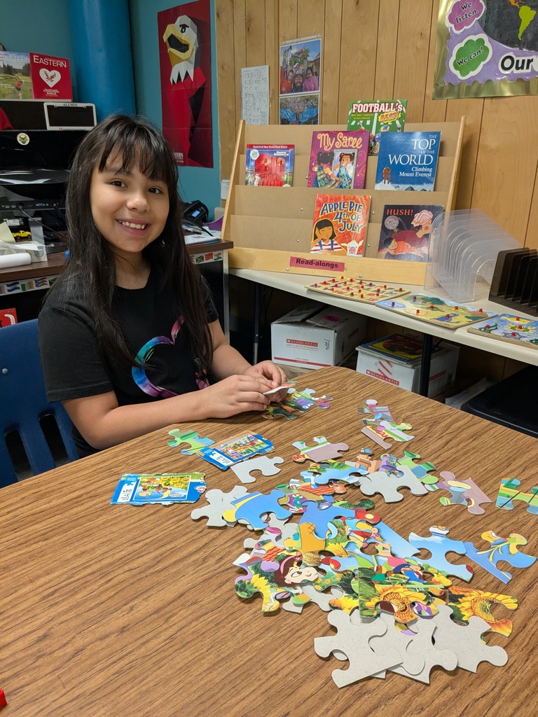 A child sits at a table working on a colorful jigsaw puzzle. Puzzle boxes and additional pieces sit nearby, with a bookshelf of children’s books in the background.