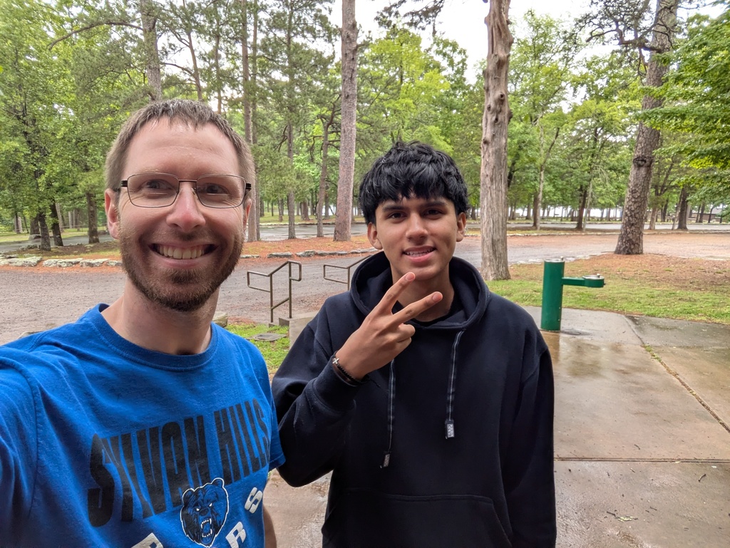 Two students stand on a paved path in a wooded park area; one wears a Sylvan Hills Bears shirt and the other makes a peace sign gesture. A green water fountain stands behind them.