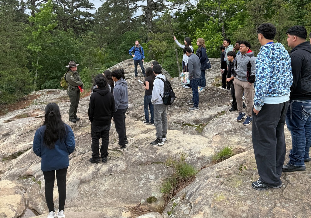 A park ranger addresses students standing on a rocky outcrop bordered by trees, with the group listening during an outdoor lesson.