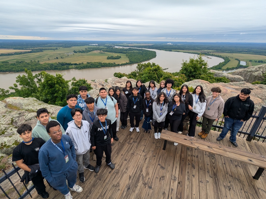 A group of students stands on a wooden observation deck overlooking a wide river that curves through green fields and forested hills under an overcast sky.