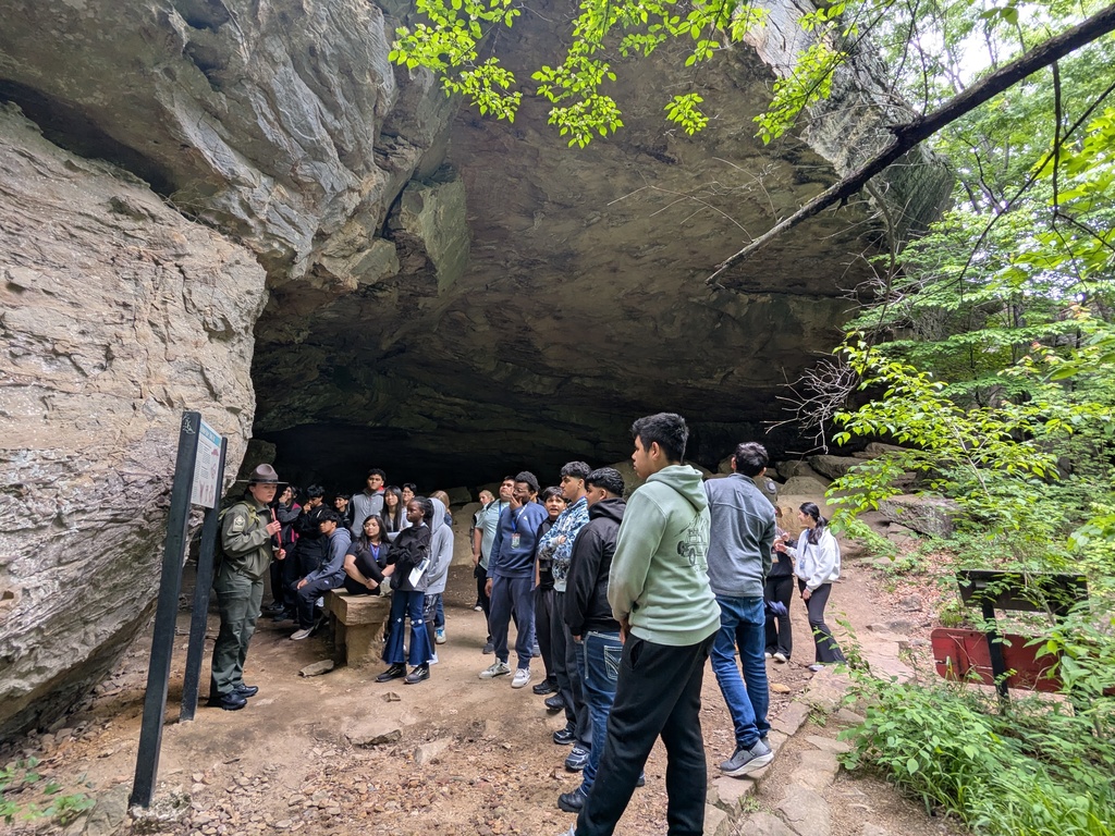 A park ranger in uniform speaks to a group of students standing at the entrance of a large rock cave surrounded by dense forest.