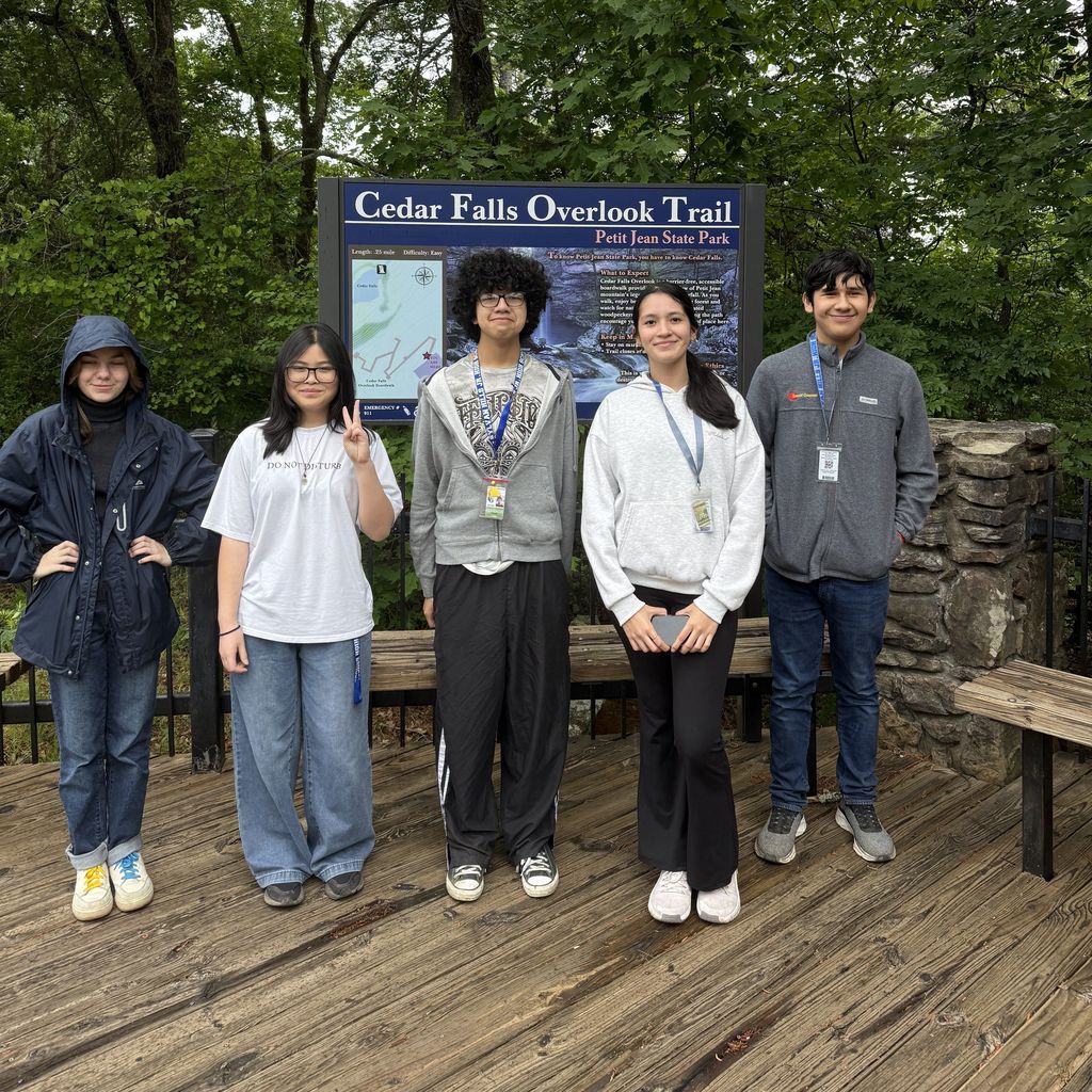 Five students stand on a wooden platform in front of the “Cedar Falls Overlook Trail” sign at Petit Jean State Park, posing for a group photo among tall trees.