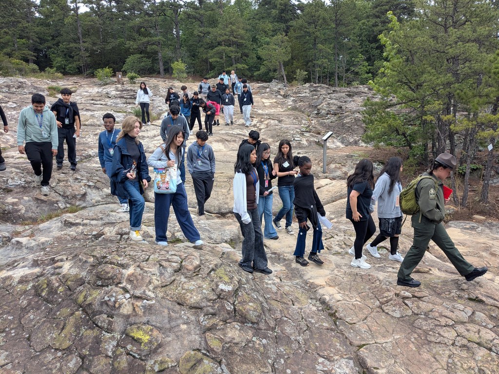A group of students walks across a broad, textured rock surface surrounded by trees while a park ranger leads them along the trail. Several students carry papers or notebooks as they explore the area.