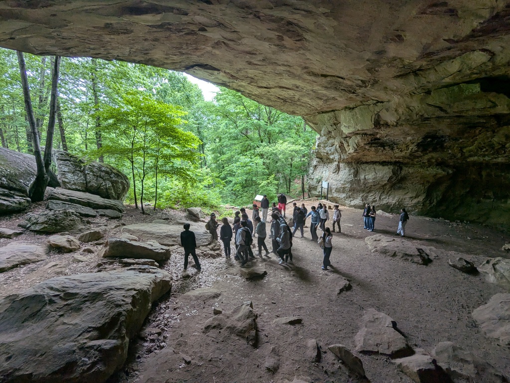 Students gather inside a large rock shelter with a wide opening and scattered boulders while trees and greenery frame the entrance.