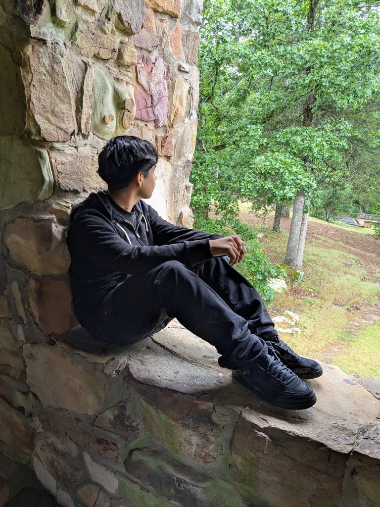 A student dressed in black sits on a built‑in stone ledge, looking out toward a wooded area with grass and trees beyond the rock wall.