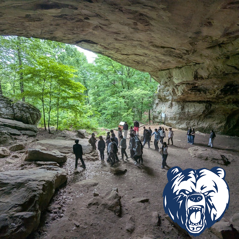 Outdoor rock shelter with students gathered under a large stone overhang during a nature lesson.