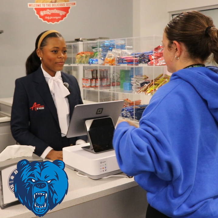 A student in an Academies of PCSSD blazer working at a cash register with another student buying something.