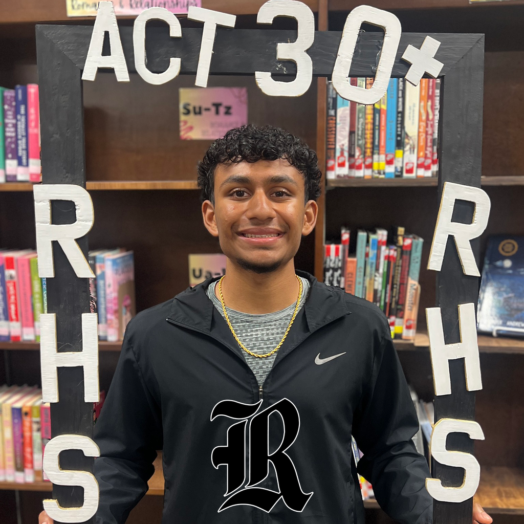 Student holding an “ACT 30+ CLUB” frame in front of library shelves.