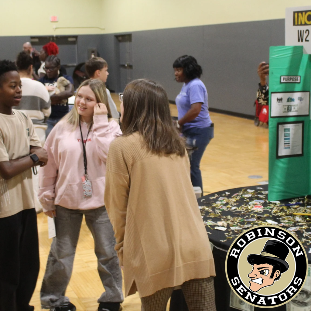 Students standing in a a gym. The ones in the foreground are listening to an adult about income taxes.