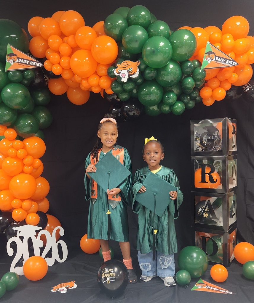 Two children in green graduation gowns hold their caps while standing in front of a balloon backdrop in orange, green, and black. The display includes Daisy Bates pennants, stacked clear boxes spelling “GRAD,” a white “2026” cutout, and a black balloon reading “Just Graduated.”