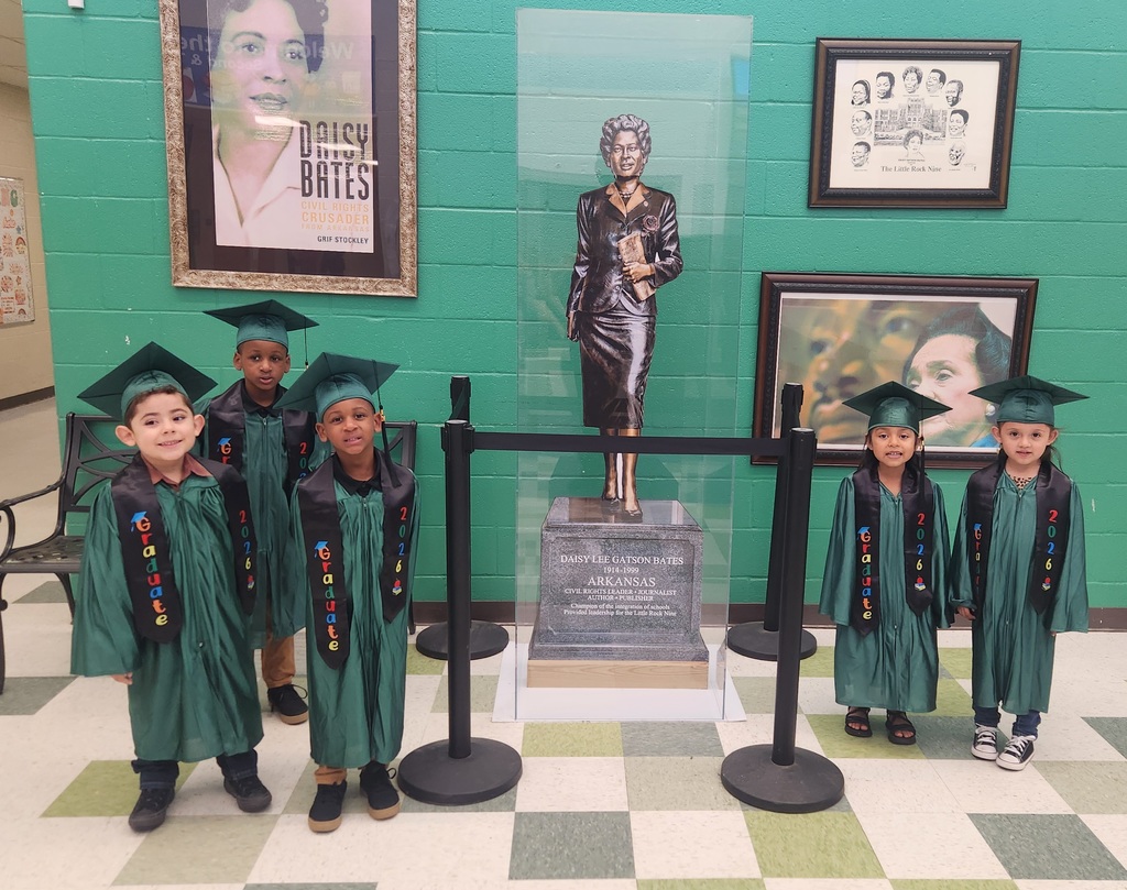 Five young children in green graduation gowns and caps stand in front of a display honoring Daisy Bates. Behind them are framed civil rights posters and a statue of Daisy Bates inside a clear case. Their stoles read “Graduate 2026.”