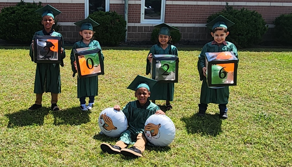 Five children in green graduation gowns and caps pose on a grassy area in front of a brick building. Four hold transparent cubes displaying the numbers “2,” “0,” “2,” and “6,” while the child seated in front holds two tiger‑face balloons.