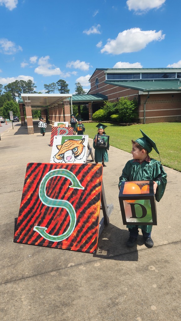 A group of children in green graduation gowns and caps stand outside a brick school building holding decorated boxes with letters, drawings, and the year “2026.” A bright blue sky with scattered clouds is visible above them.