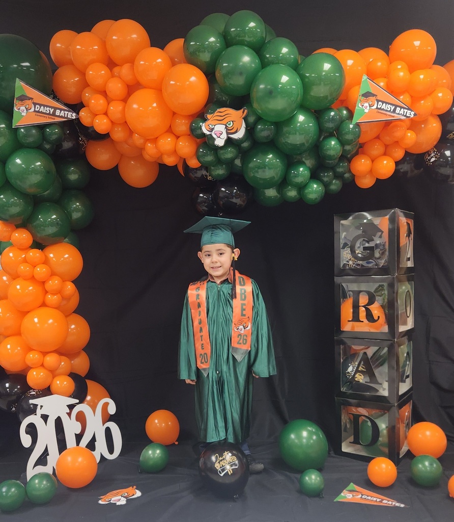 A young graduate wearing a green gown and cap with orange “GRADUATE 2026” and “DBE” stoles stands in front of a balloon arch in orange, green, and black. The backdrop includes Daisy Bates pennants, stacked “GRAD” boxes, a white “2026” cutout, and a black “Just Graduated” balloon.
