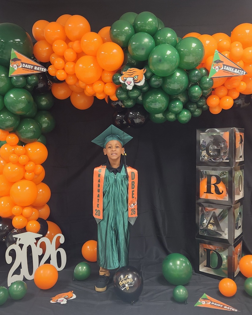 A young graduate wearing a green gown and cap with orange “GRADUATE 2026” and “DBE” stoles stands in front of a balloon arch in orange, green, and black. The backdrop includes Daisy Bates pennants, stacked “GRAD” boxes, a white “2026” cutout, and a black “Just Graduated” balloon.