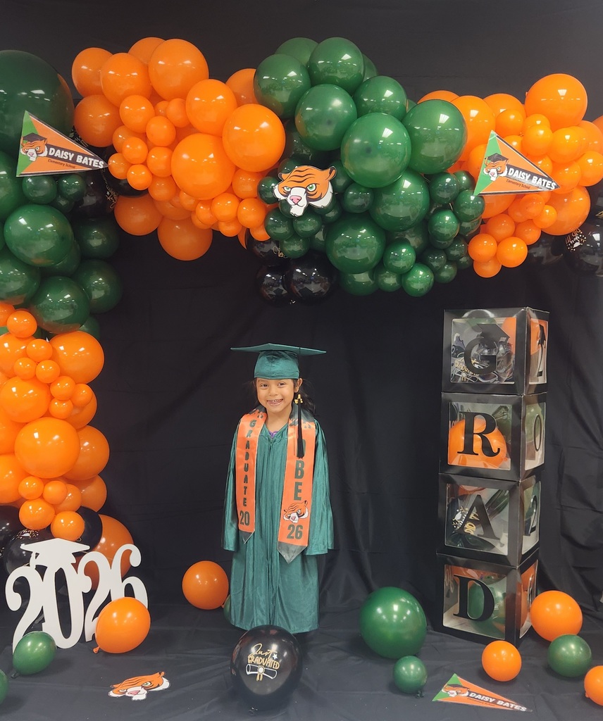 A young graduate wearing a green gown and cap with orange “GRADUATE 2026” and “DBE” stoles stands in front of a balloon arch in orange, green, and black. The backdrop includes Daisy Bates pennants, stacked “GRAD” boxes, a white “2026” cutout, and a black “Just Graduated” balloon.
