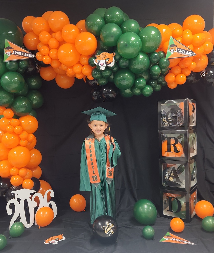 A young graduate wearing a green gown and cap with orange “GRADUATE 2026” and “DBE” stoles stands in front of a balloon arch in orange, green, and black. The backdrop includes Daisy Bates pennants, stacked “GRAD” boxes, a white “2026” cutout, and a black “Just Graduated” balloon.