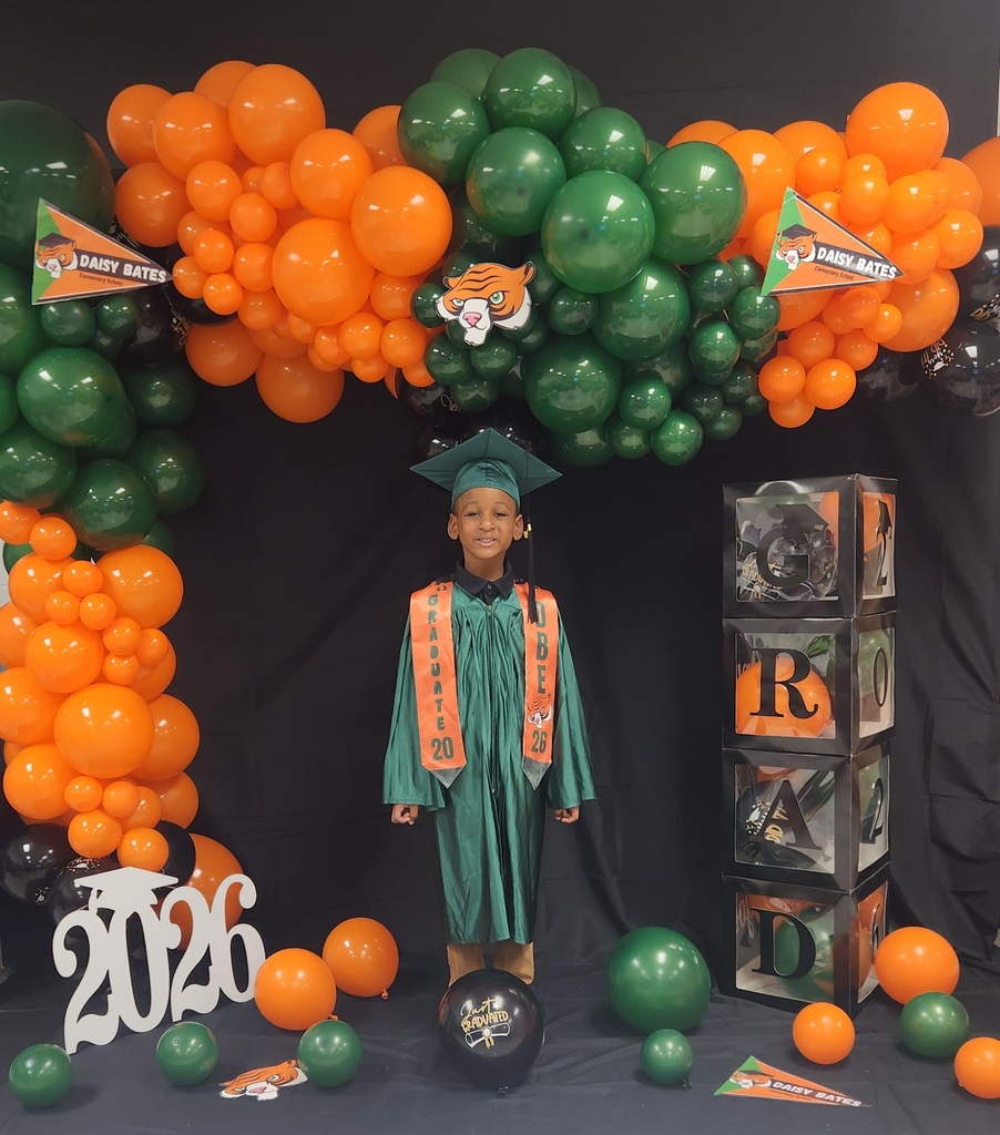 A young graduate wearing a green gown and cap with orange “GRADUATE 2026” and “DBE” stoles stands in front of a balloon arch in orange, green, and black. The backdrop includes Daisy Bates pennants, stacked “GRAD” boxes, a white “2026” cutout, and a black “Just Graduated” balloon.