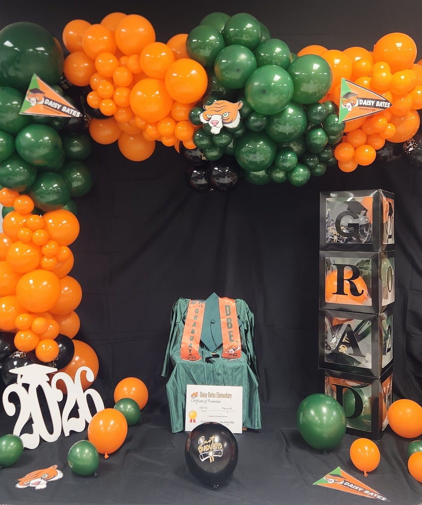 A graduation display features a green gown with orange “GRADUATE 2026” and “DBE” stoles, surrounded by orange, green, and black balloons. A “2026” sign sits on the floor next to stacked clear boxes spelling “GRAD,” Daisy Bates pennants, and a promotion certificate.
