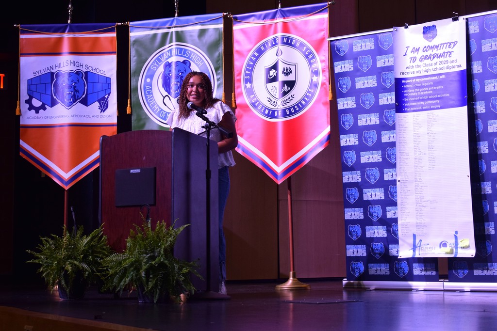 A speaker stands at a podium in front of three Sylvan Hills High School academy banners representing Engineering, Medicine, and Business.