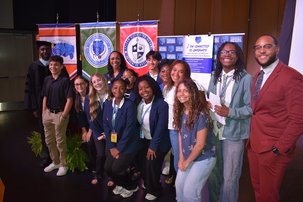A group of students and educators stand together onstage in front of three Sylvan Hills High School academy banners and a large “I Am Committed to Graduate” pledge poster.