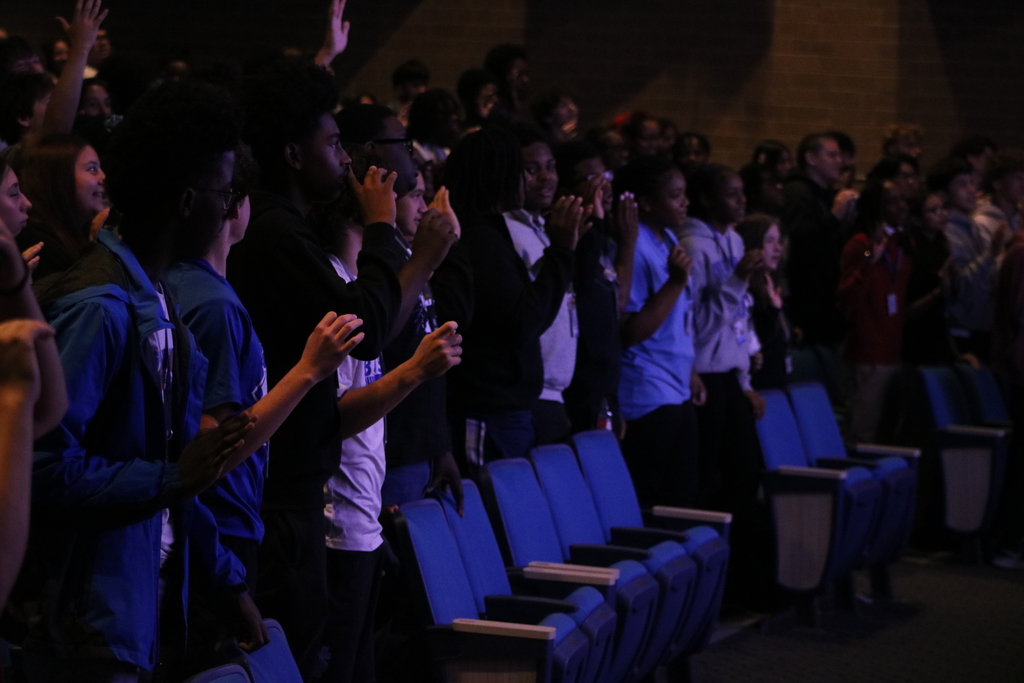 A large audience stands in an auditorium with blue seats, many raising their hands or clapping during an event.