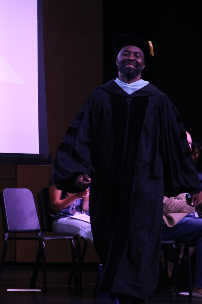 A person in full academic regalia—black doctoral gown, velvet panels, and a tam with a gold tassel—stands onstage during a ceremony, with seated attendees and purple lighting in the background.