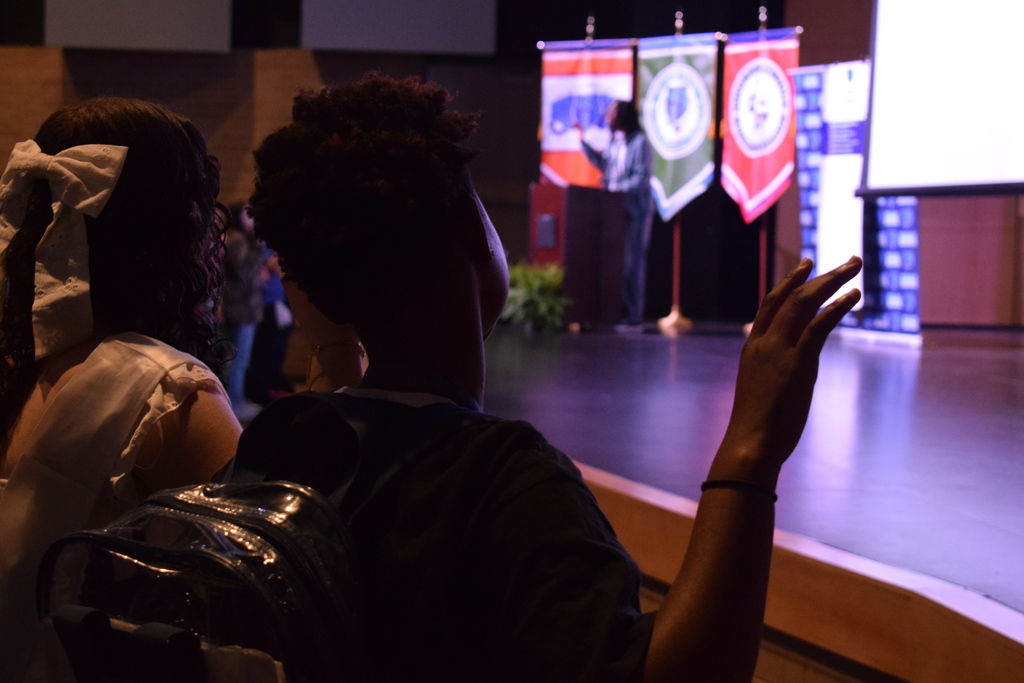 Two students stand near the front of the auditorium, facing the stage where a presenter speaks at a podium. Several colorful academy banners hang behind the speaker, and a projection screen is visible to the right.