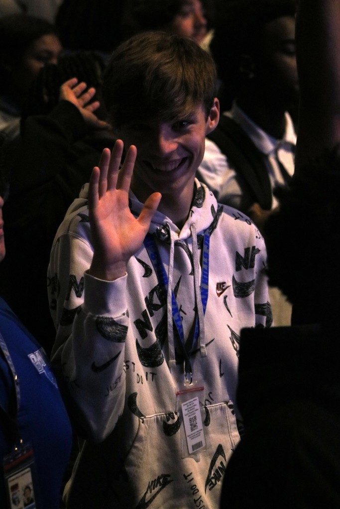 A student wearing a white Nike “Just Do It” hoodie and a blue lanyard raises a hand in a wave while standing among a group of people in a dimly lit auditorium.