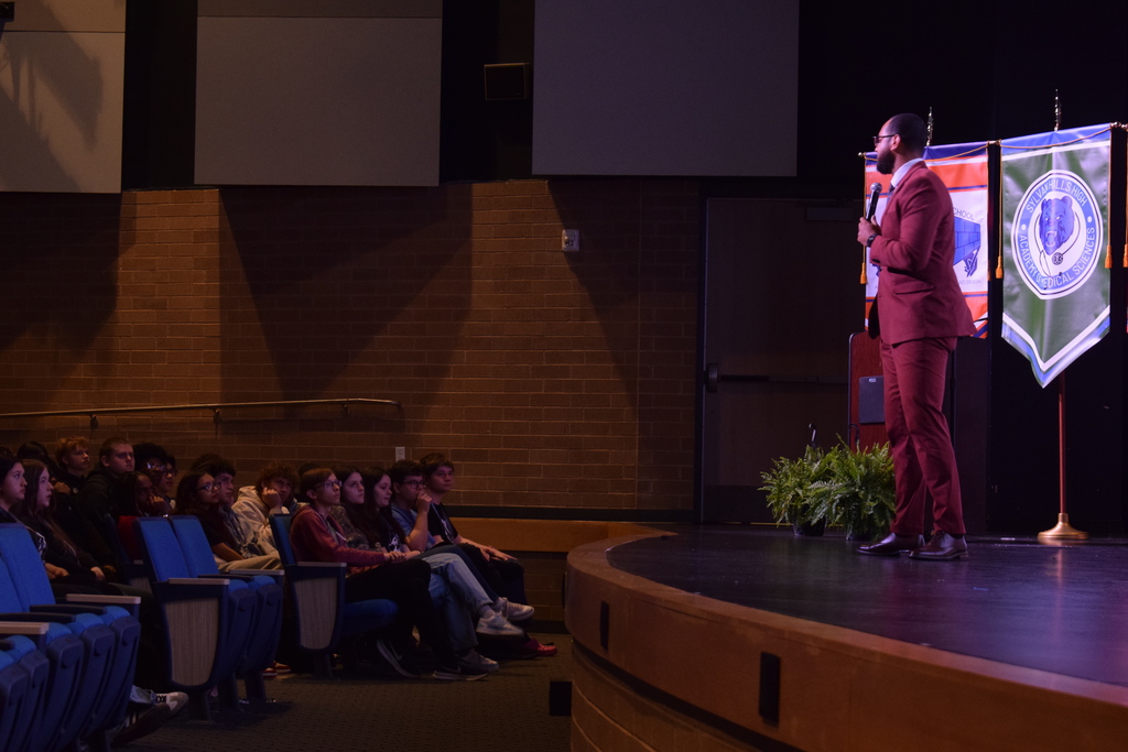 A speaker in a maroon suit stands onstage at the Sylvan Hills High School Performing Arts Center, addressing students seated in blue auditorium chairs. Two academy banners hang behind the speaker.