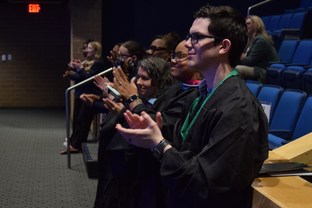 Graduates in black gowns sit in rows of blue auditorium seats, clapping toward the stage during a ceremony.